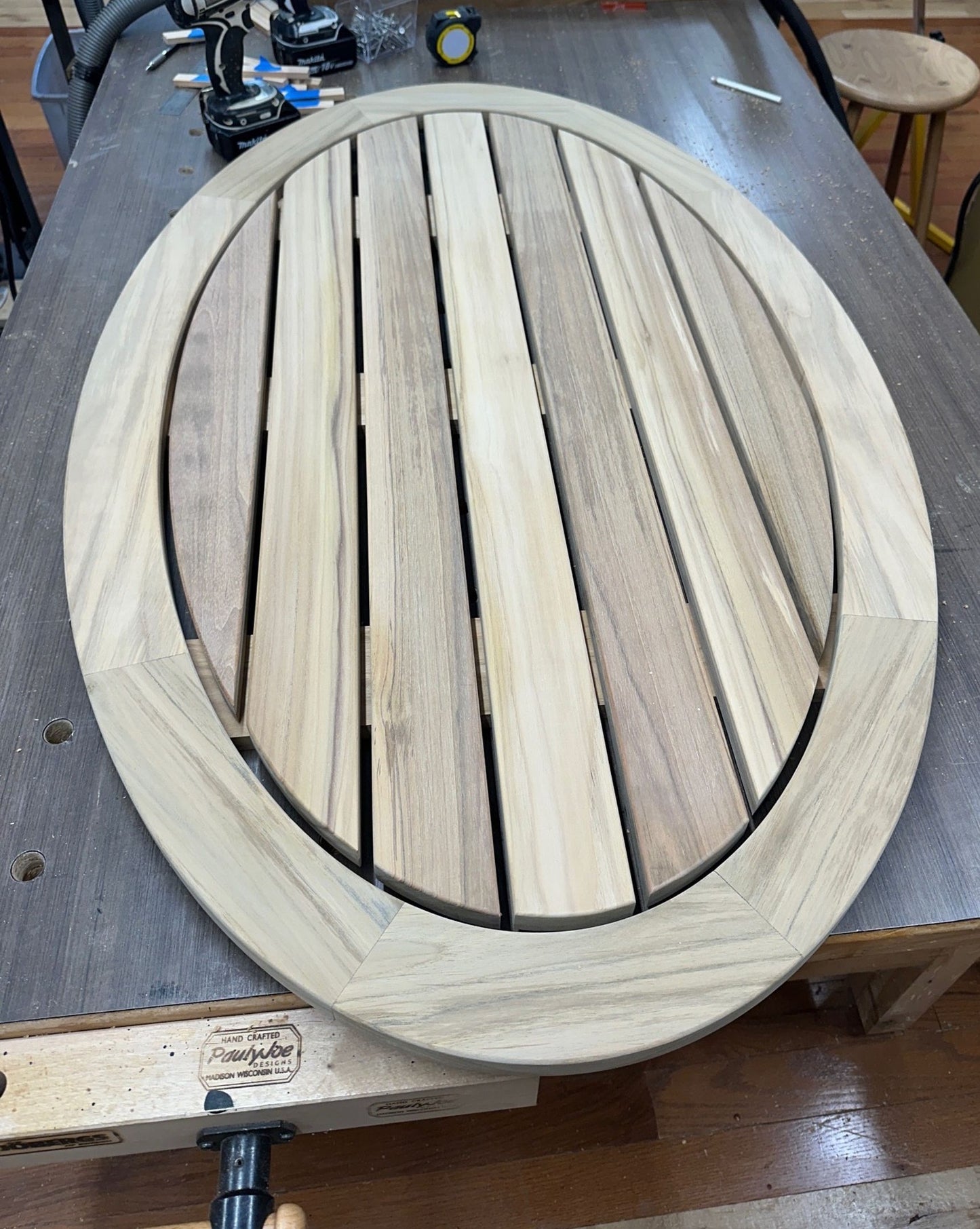 An oval wooden slatted table top or deck surface with a thick round border, photographed on a workbench in a workshop setting.