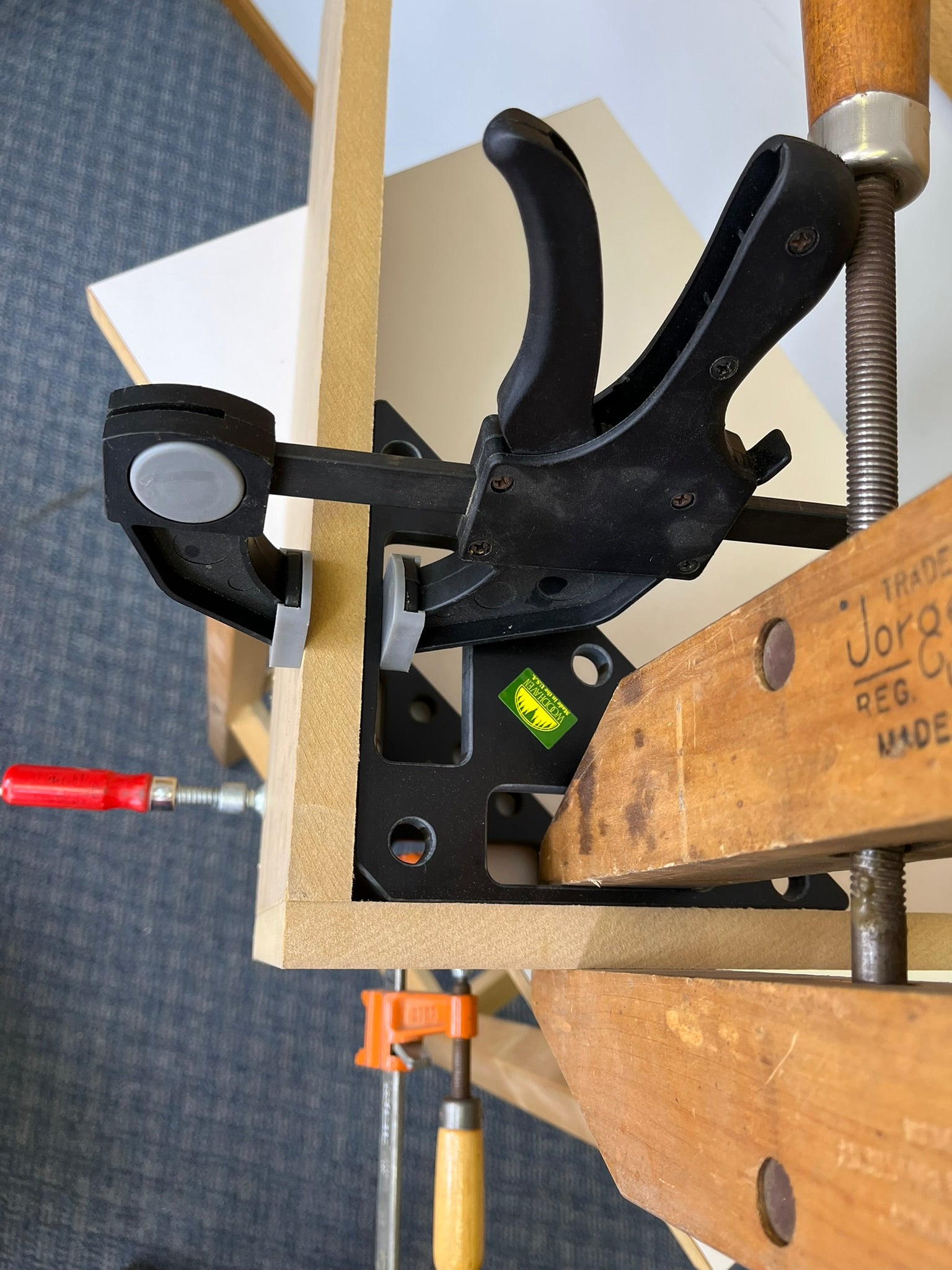 A close-up of a black metal clamp holding a wooden frame or jig in place. The setup is photographed in a workshop environment.