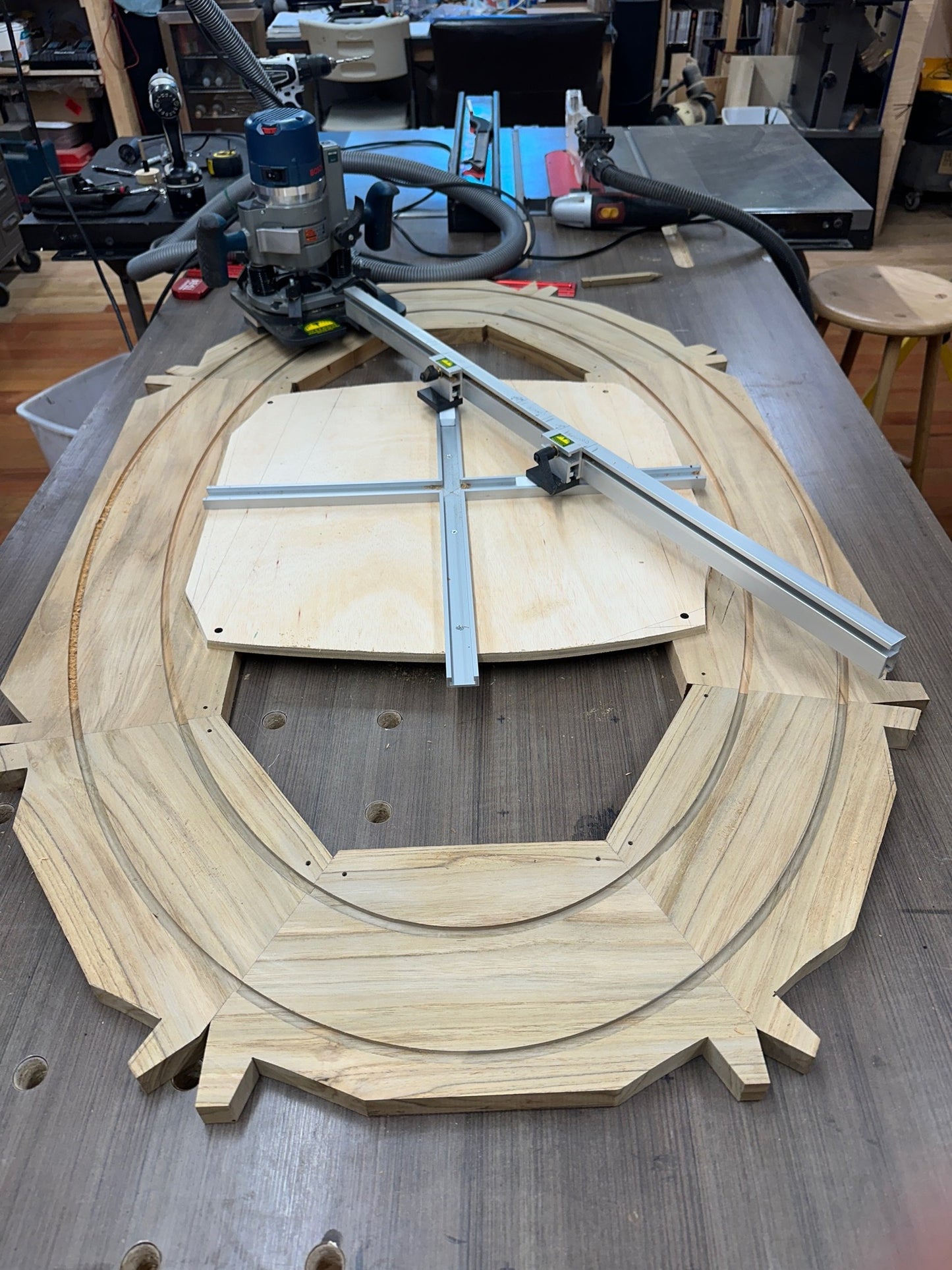 A large unfinished wooden oval table shown on a workbench in a workshop. A circular router with a guide rail and a plunge router are positioned above it, a woodworking project in progress.