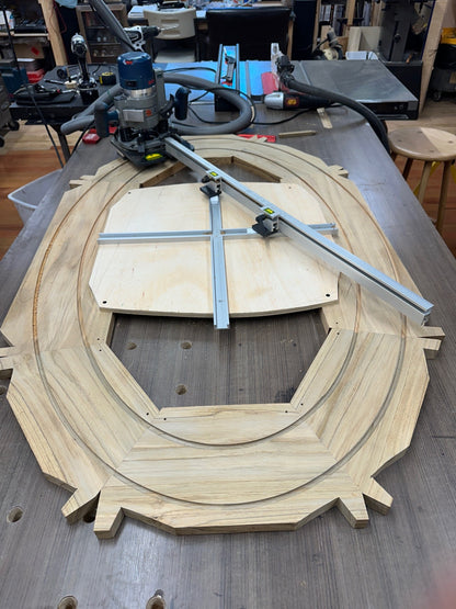 A large unfinished wooden oval table shown on a workbench in a workshop. A circular router with a guide rail and a plunge router are positioned above it, a woodworking project in progress.