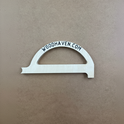 A light-colored wooden decorative piece with a semi-circular top and the text 'WOODHAVEN.COM' printed in black along the curve, placed on a textured brown background.